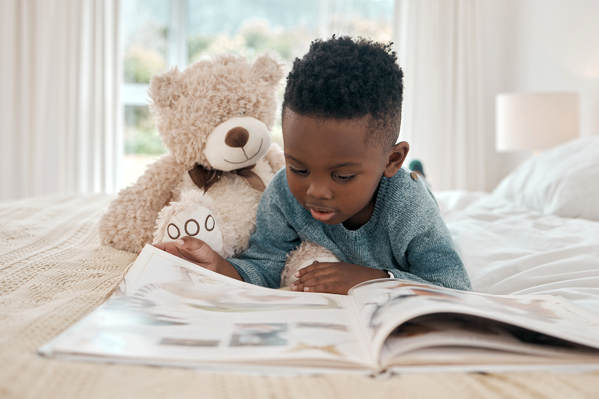 A young boy lying on a bed, looking at an open photo album, with a stuffed teddy bear beside him in a bright bedroom.
