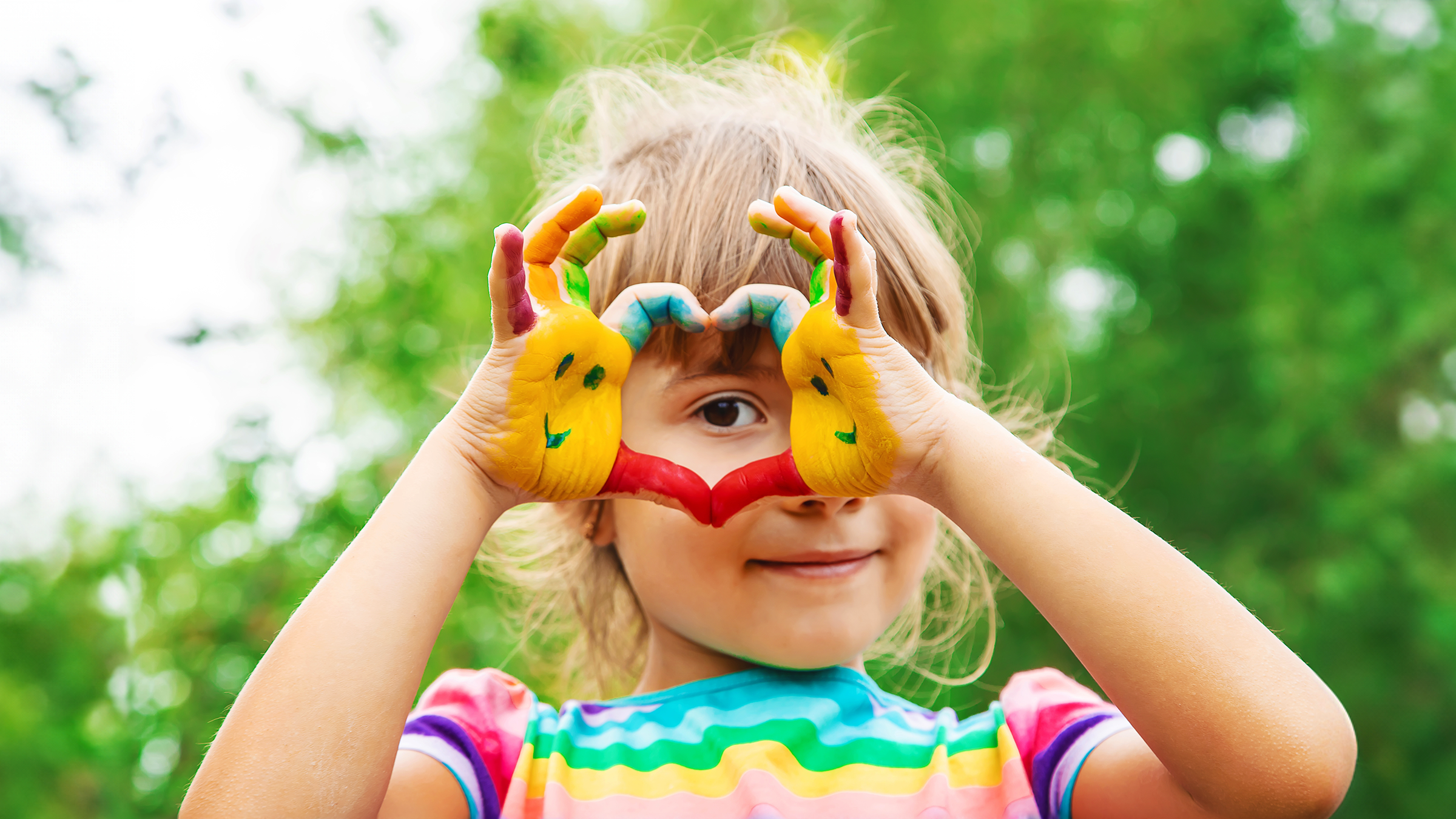 A young girl with painted hands holding a heart-shaped gesture around her eye outdoors.