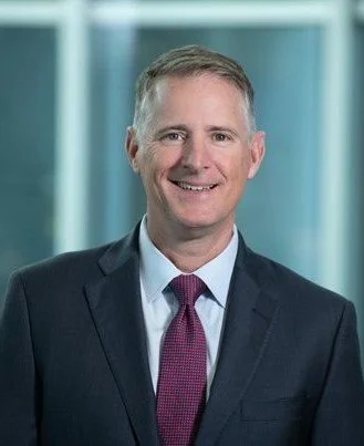 Professional man in a dark suit, white shirt, and red tie, smiling in front of a glass building background.