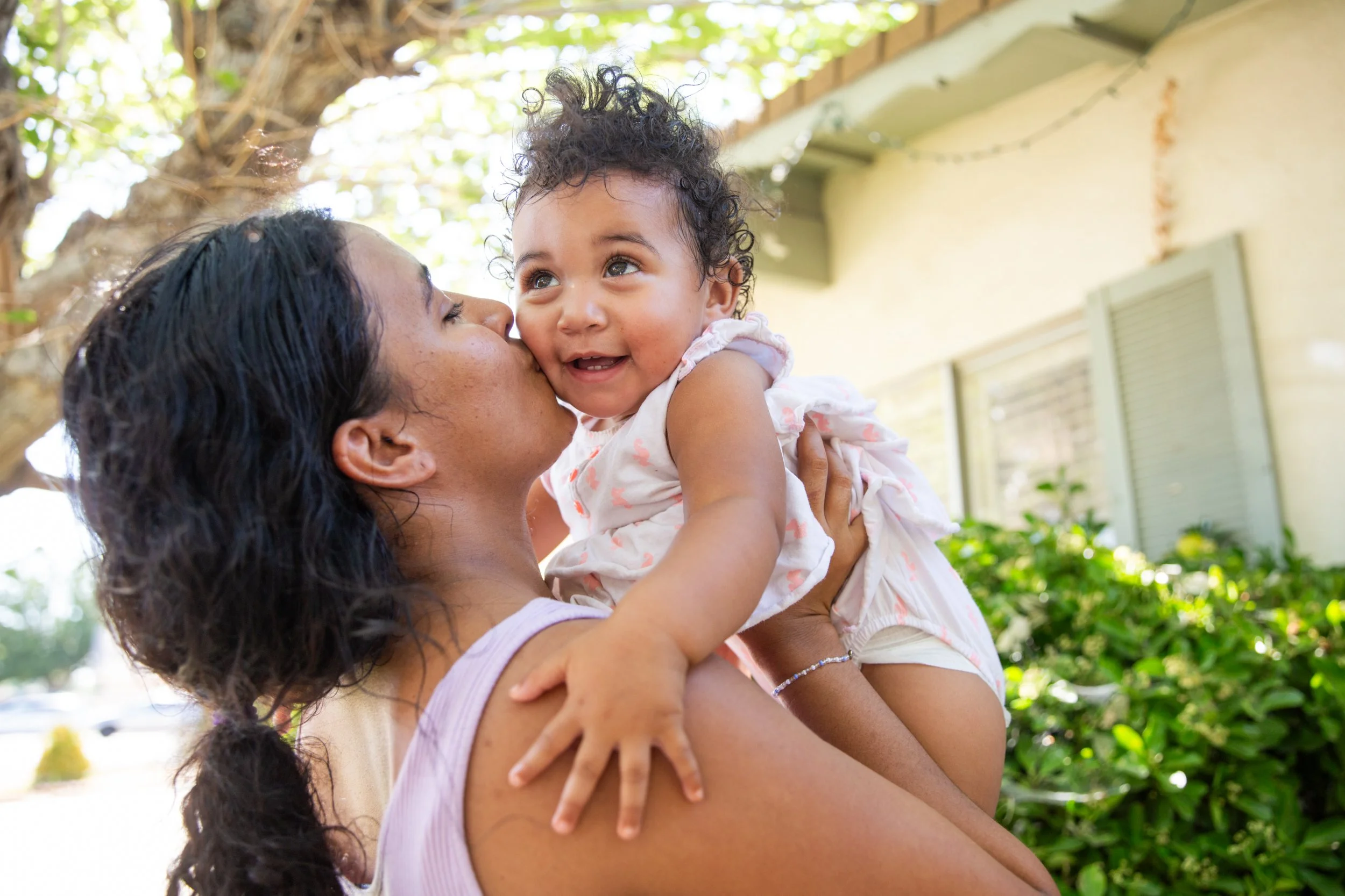 A woman is holding a young girl up in the air outdoors, with trees and a house in the background. The girl is smiling and looking at the woman, who is kissing her on the cheek.