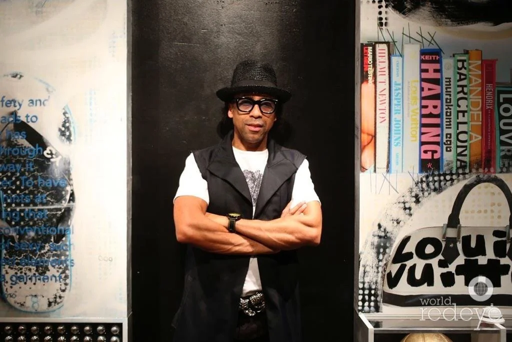 A man wearing a black hat, glasses, a black vest over a white t-shirt, and a watch, standing with arms crossed in front of a wall with books and artwork.