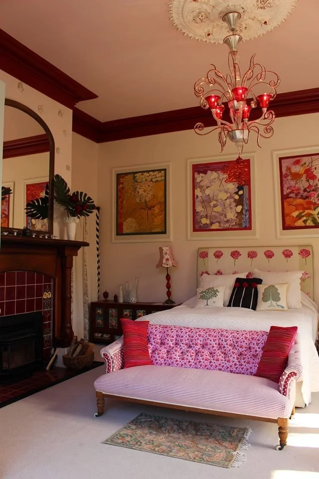 A cozy bedroom with a white bed, floral headboard, and decorative pillows. Behind the bed are three framed botanical artwork pieces. There is a pink and red floral-patterned sofa at the foot of the bed. A vintage side table with a lamp, a fireplace w