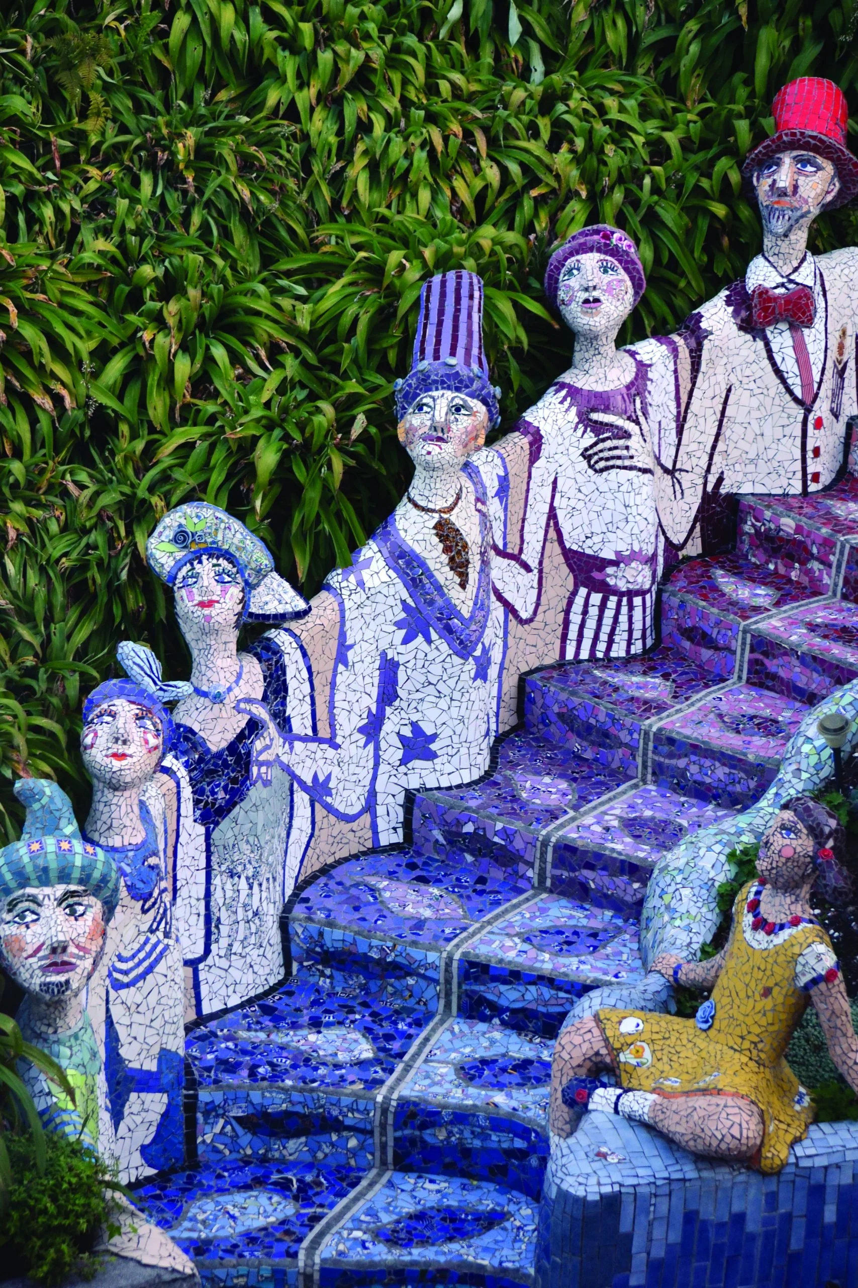 Colorful mosaic sculpture of a family with seven members, sitting on a blue mosaic bench, surrounded by green foliage.