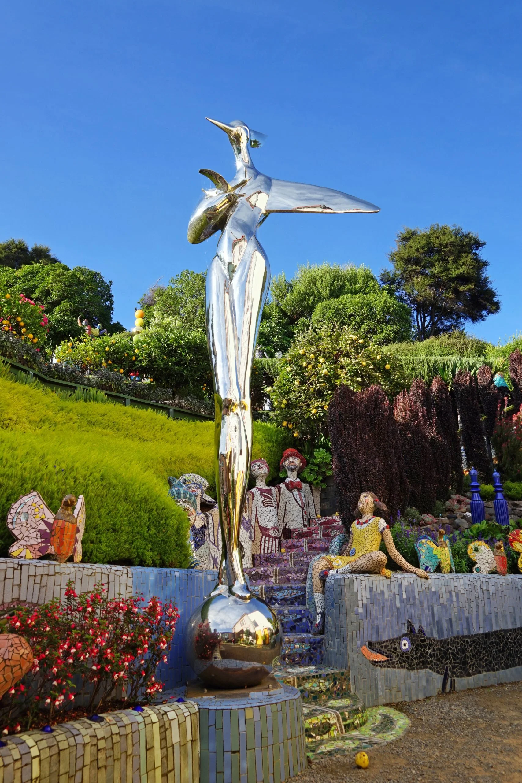 Shiny metallic sculpture of a woman holding a whale, surrounded by mosaic art and colorful plants in a garden setting.