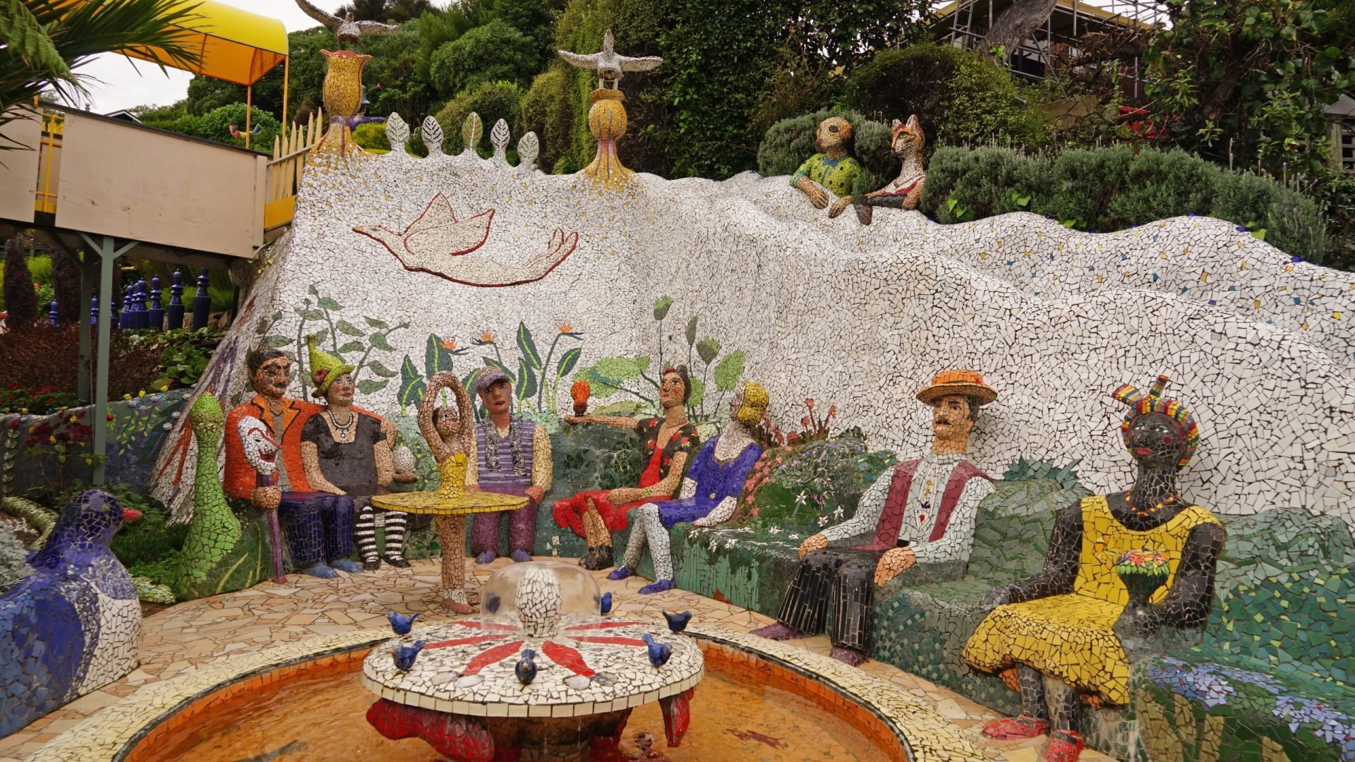 Colorful mosaic sculpture of people sitting around a fountain, with additional mosaic animals and birds, and lush greenery in the background.
