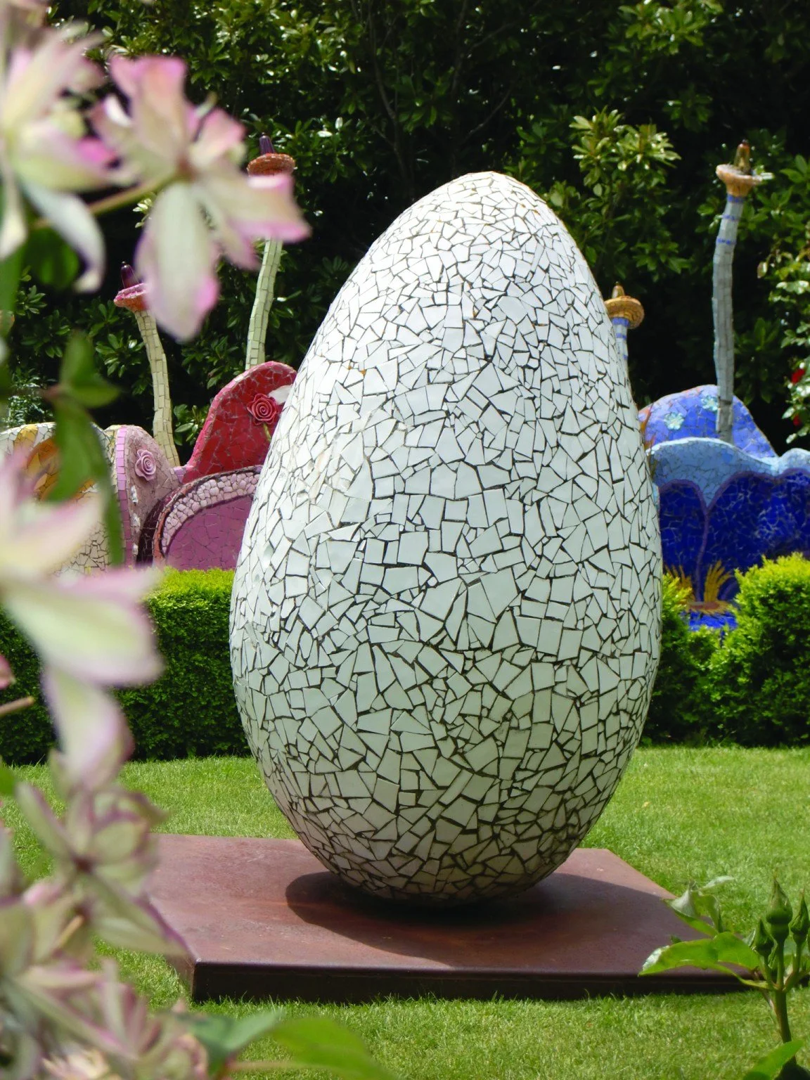 Large mosaic sculpture of an egg with a black and white crackle pattern, placed on a red platform in a garden with greenery and colorful abstract structures in the background.
