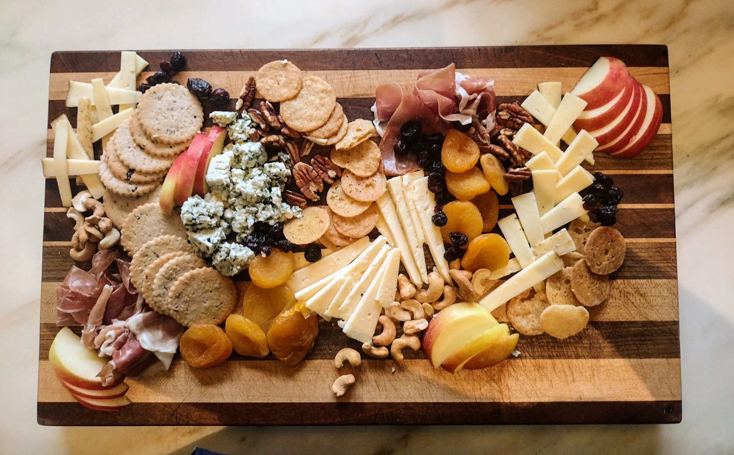 A charcuterie board with an assortment of cheese, crackers, dried apricots, apple slices, nuts, and cured meats on a wooden cutting board.