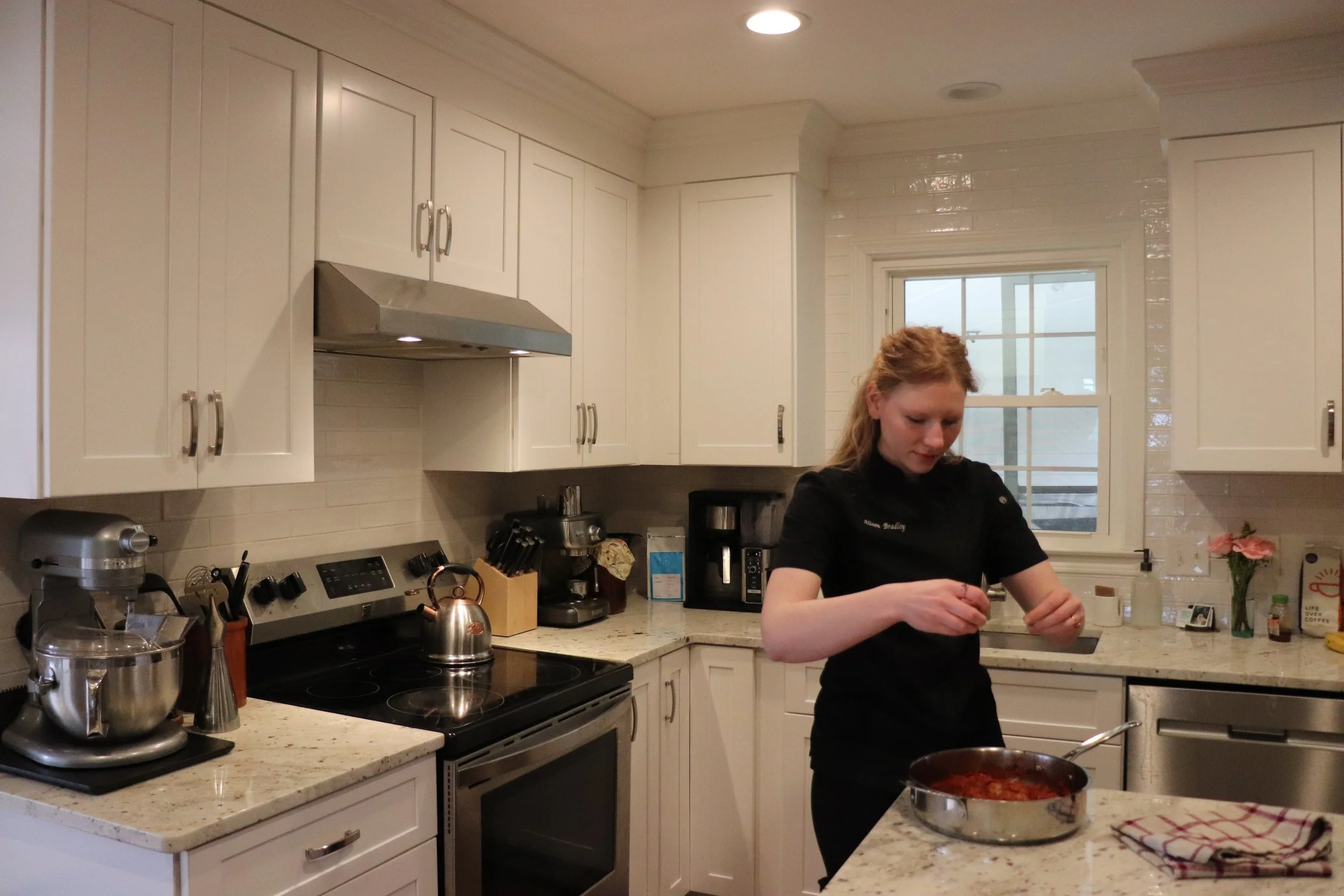 A woman in a black chef coat cooking in a white kitchen, standing near a countertop with a pot of sauce, with various appliances and kitchen items around.