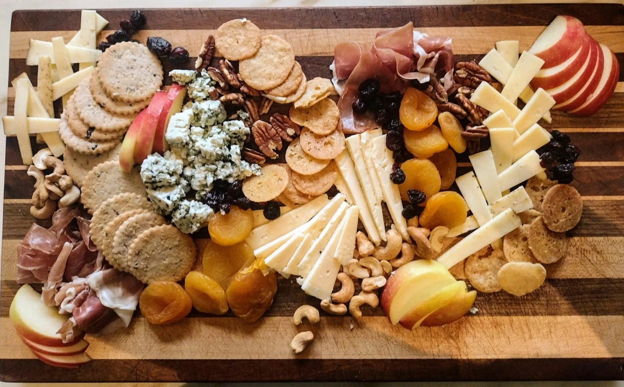 Assorted cheese, crackers, sliced apples, grapes, dried apricots, nuts, and cured meats arranged on a wooden cutting board.