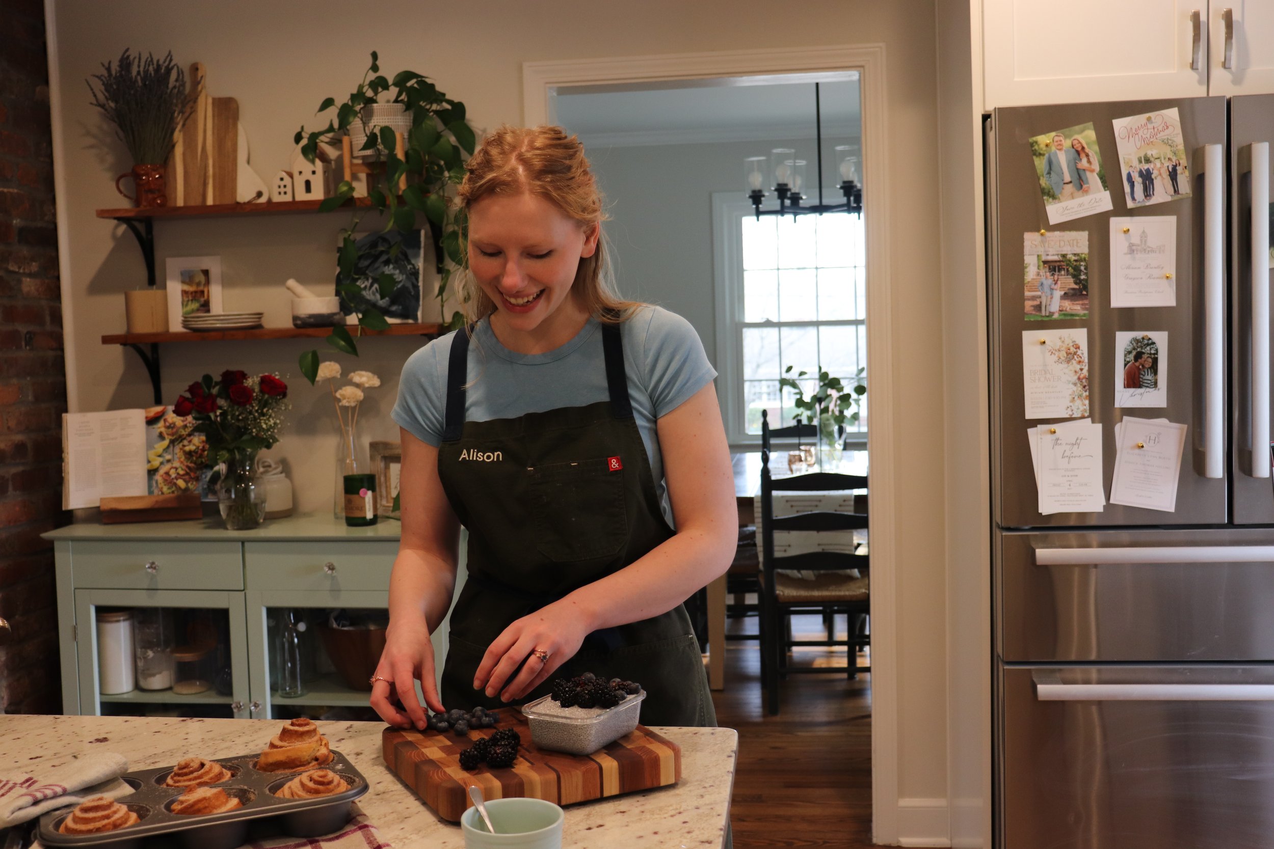 A woman with red hair, wearing a black apron with the name "Alison", smiling while preparing blackberries on a wooden cutting board in a kitchen.