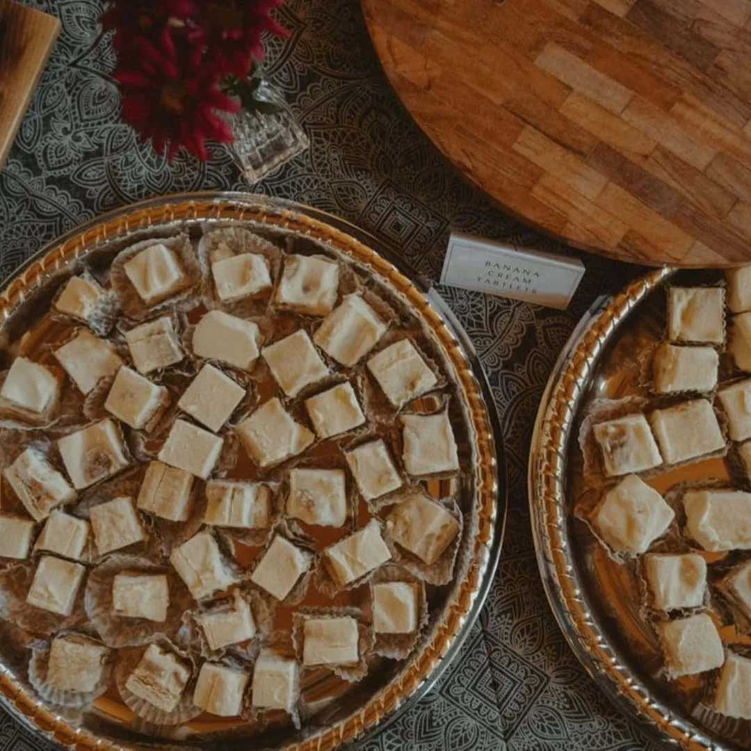 Two metal trays filled with small pieces of banana cake, placed on a patterned cloth. A small card with the words "BANANA CAKE" is visible near the trays, and a wooden cutting board is in the background.