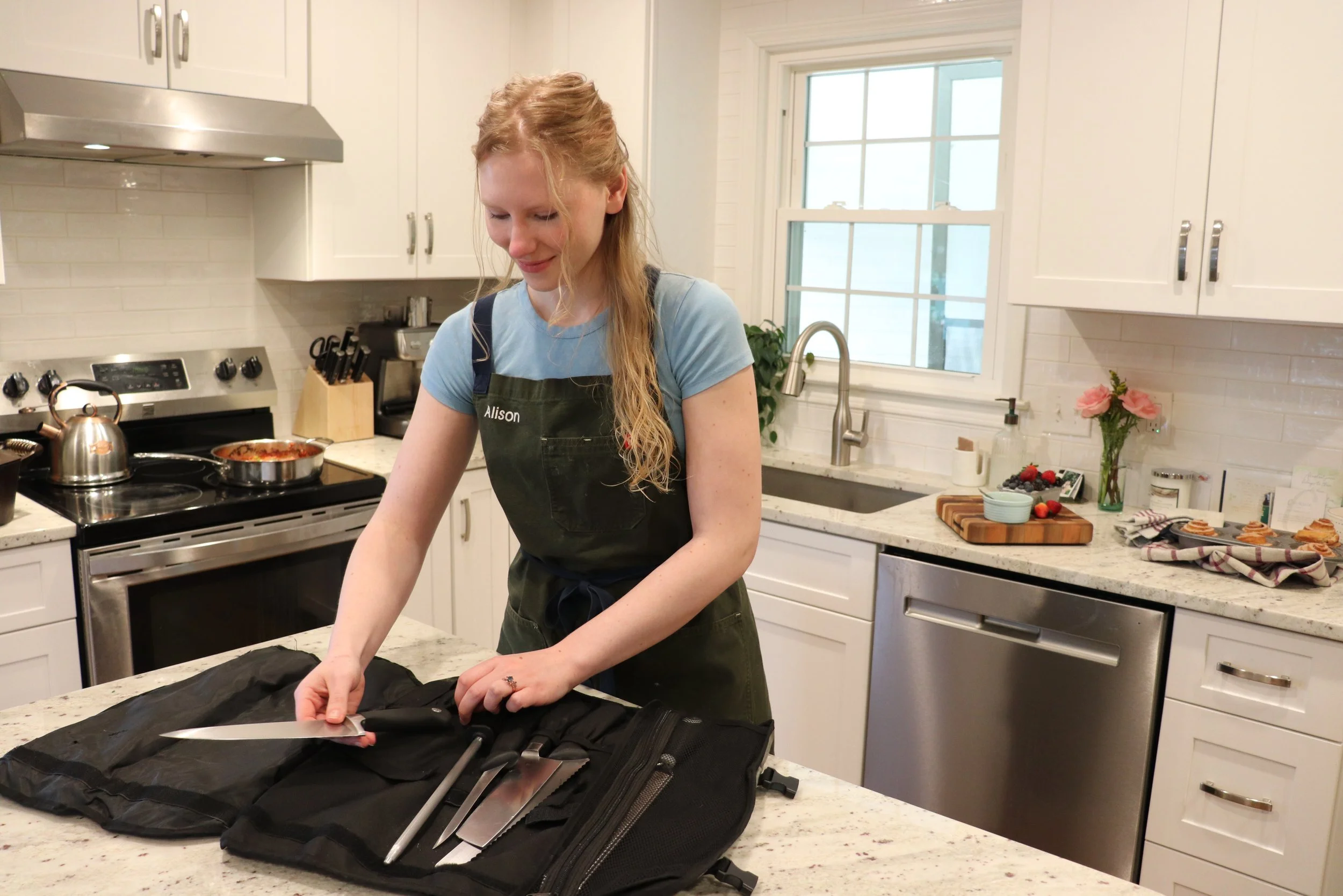 A young woman with long blonde hair wearing a green apron labeled 'Alison' is preparing knives and utensils from a black bag on a kitchen counter.