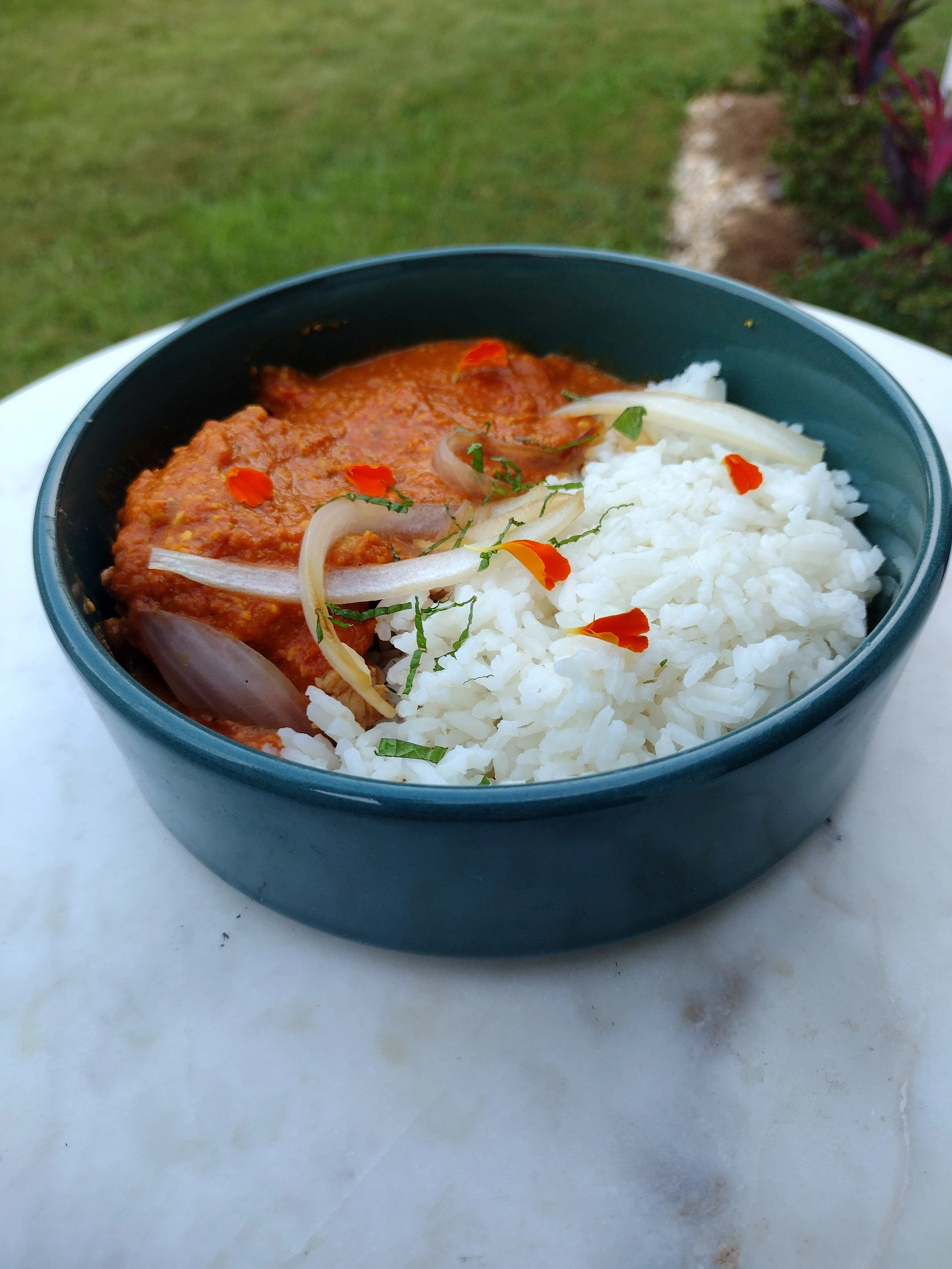 A bowl of Indian food with rice and chicken curry garnished with sliced onions and red chili flakes outside on a white surface.
