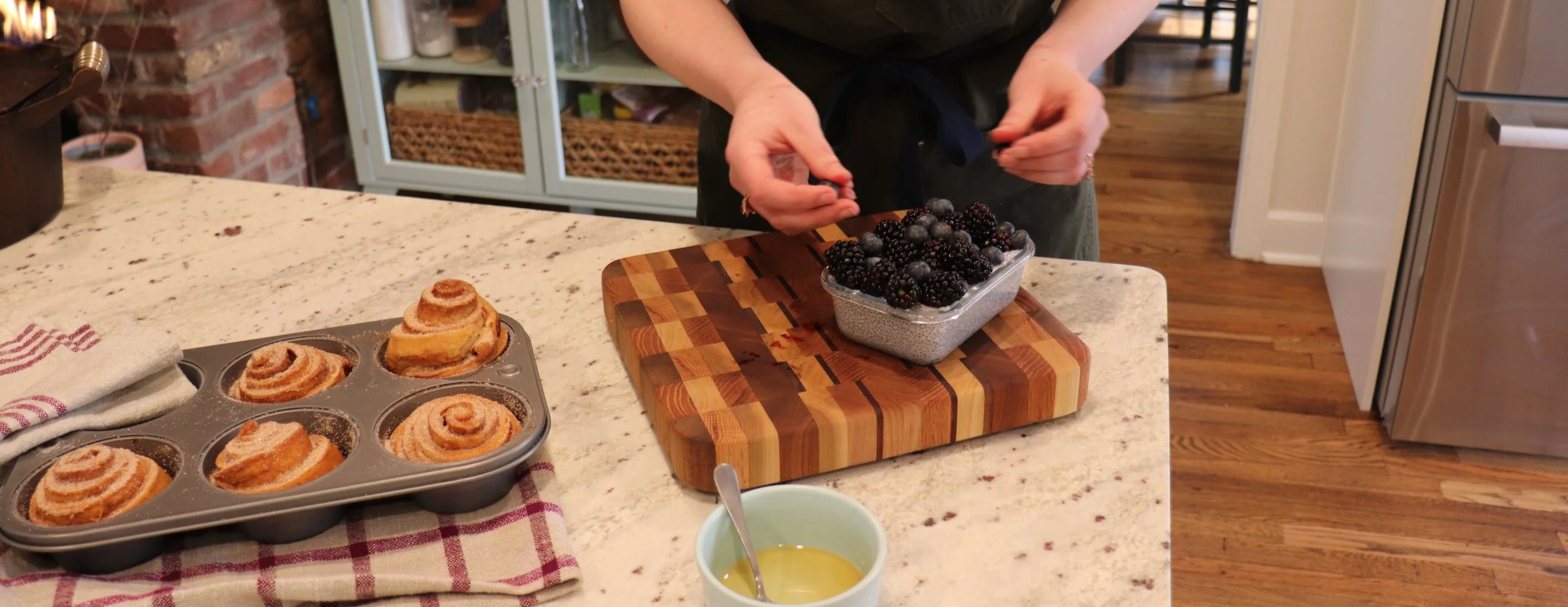 A person is preparing blackberries on a wooden cutting board in a kitchen. There are cinnamon rolls in a muffin tin and a cup of melted butter on the counter.