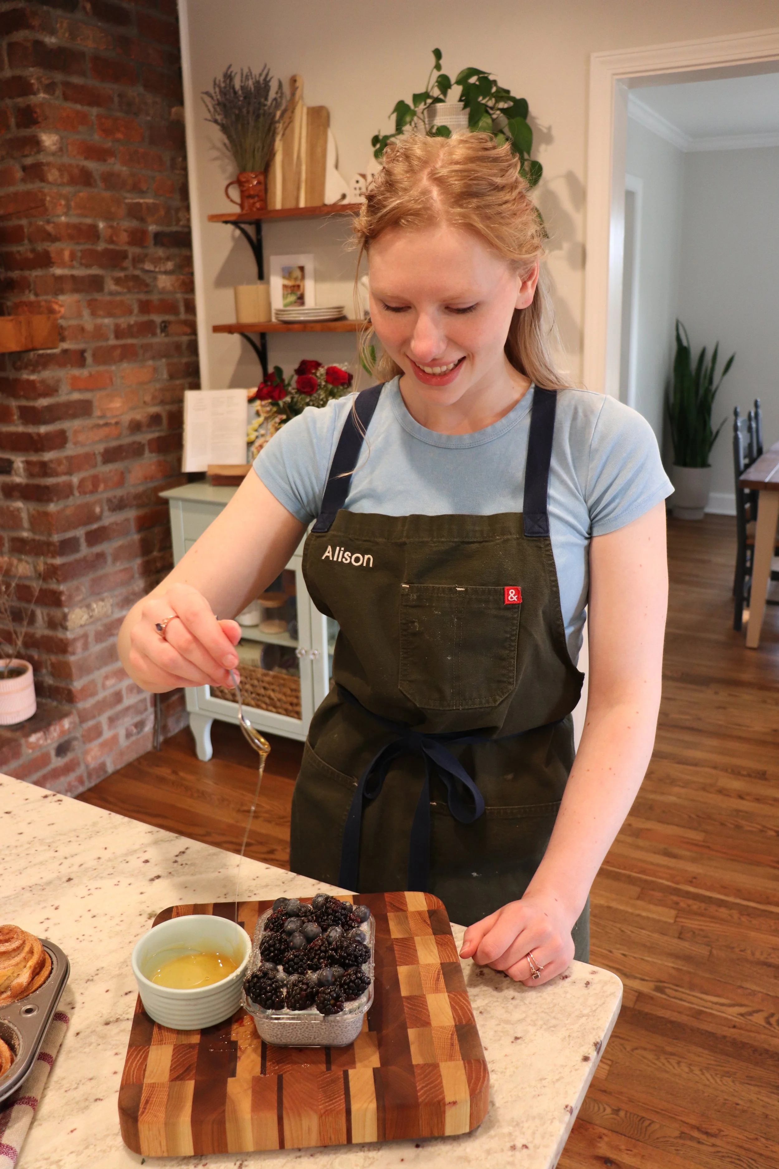 A woman with red hair, wearing a blue shirt and an apron with the name 'Alison' on it, is smiling and drizzling honey over a container of blackberries on a wooden cutting board in a cozy kitchen.