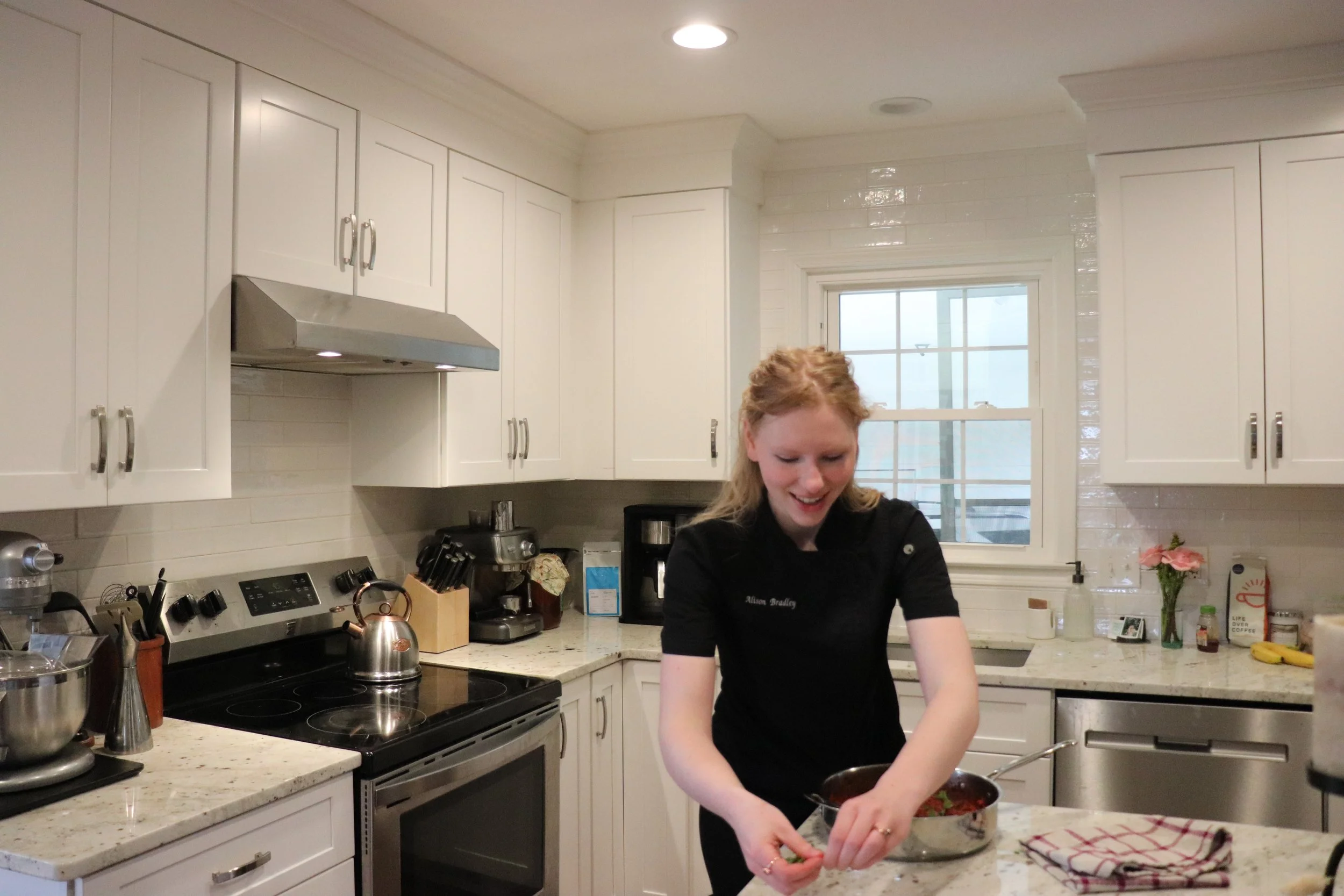 A woman with blonde hair smiling and cooking in a bright, white kitchen with white cabinets and a window in the background.