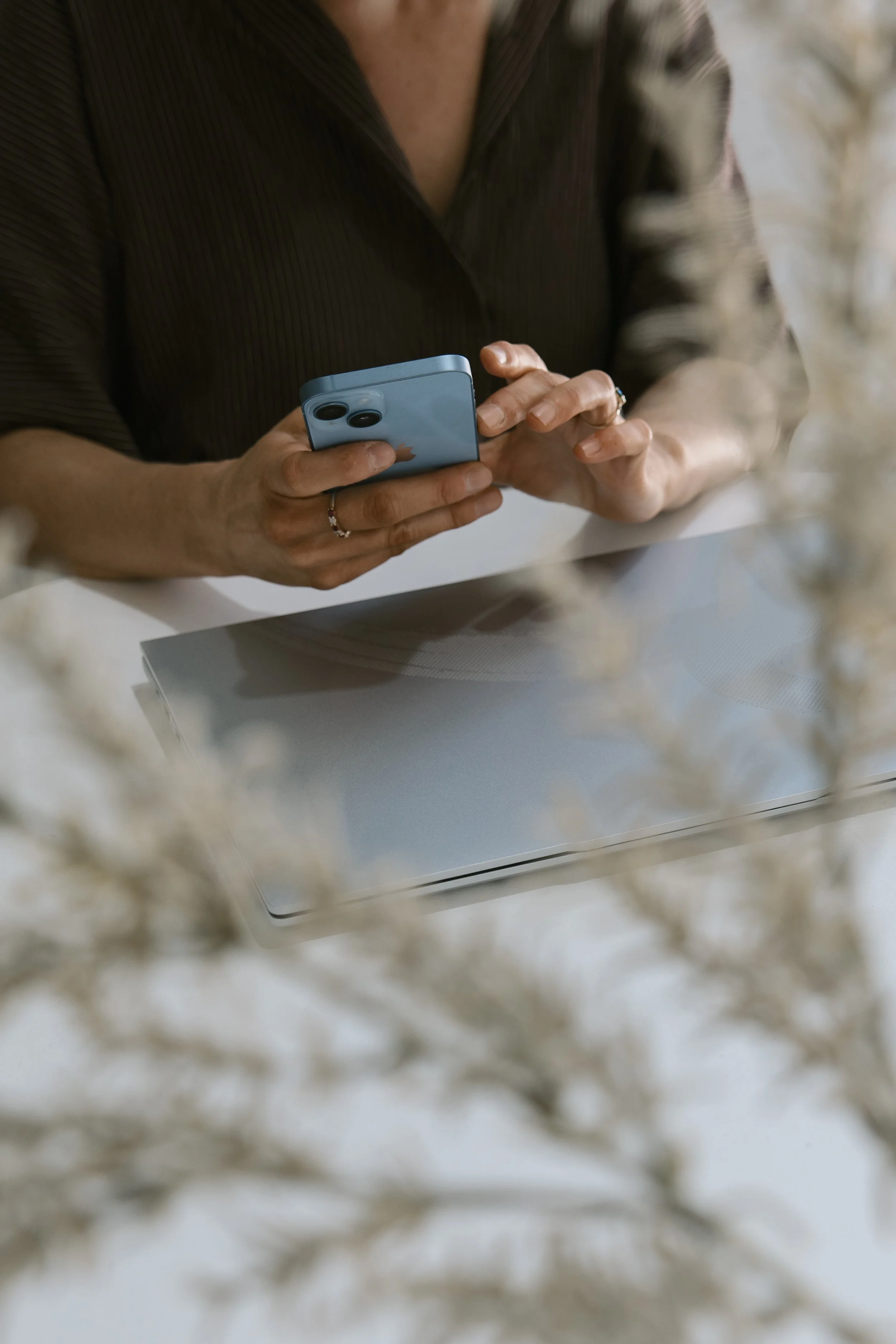 A person using a smartphone at a desk with a closed laptop, with blurred dried flowers in the foreground.