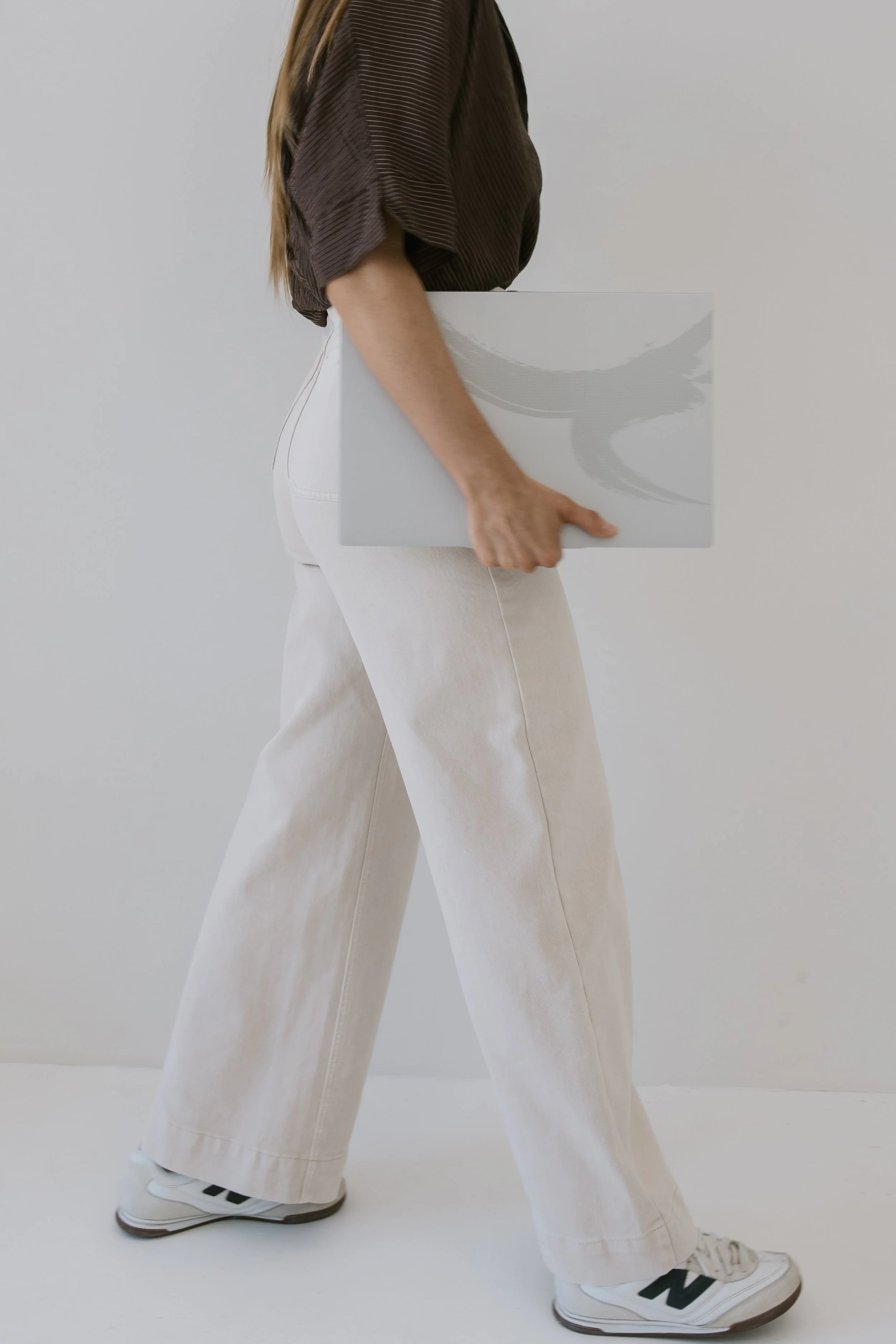 Person walking with a laptop in hand, wearing beige wide-leg pants, white sneakers, and a brown top, against a plain light background.