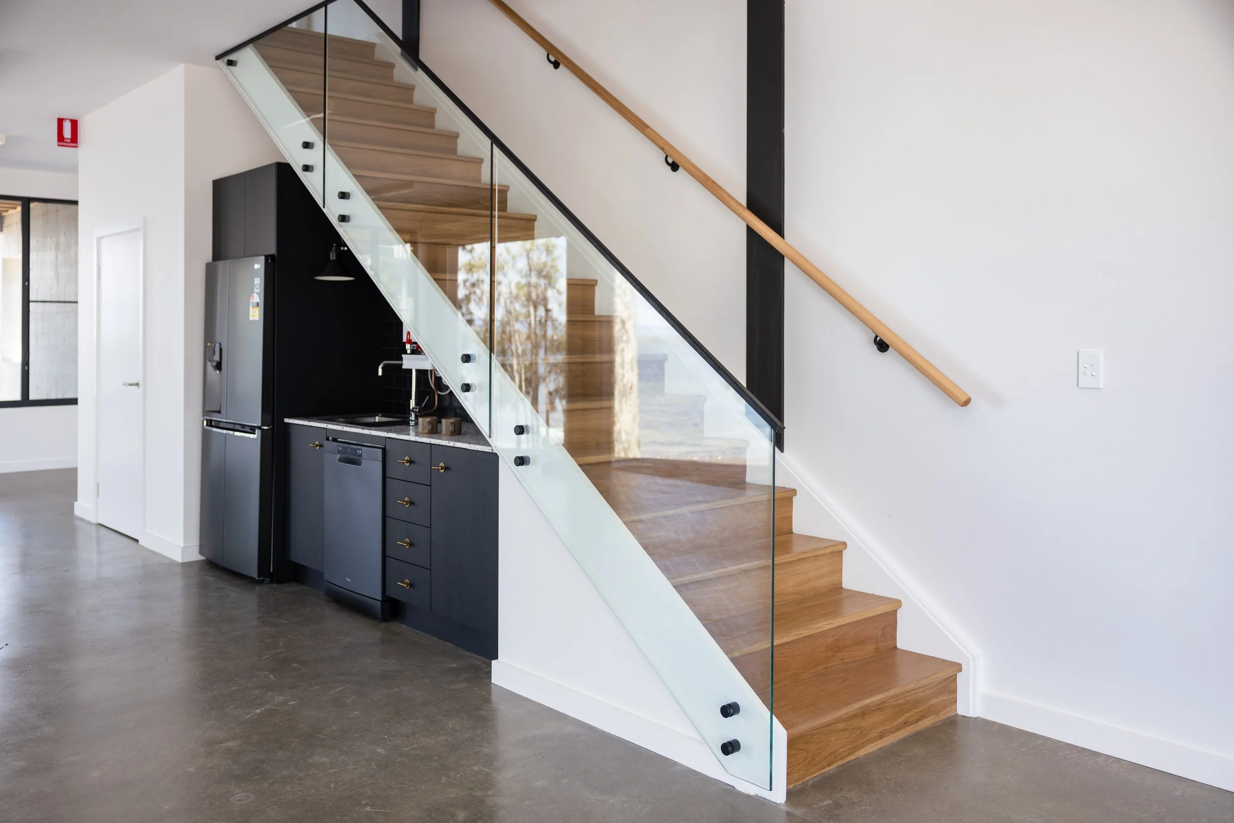 Interior view of a modern home featuring a staircase with wooden steps, glass railing, and white wall, adjacent to a black kitchen with a stainless steel refrigerator and black cabinets, amid an open floor plan with polished concrete flooring.