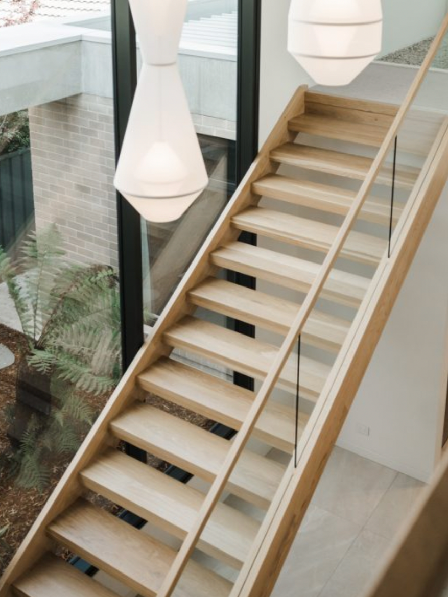 Wooden staircase with glass railing next to large windows, modern pendant lights hanging above, and a view of an indoor garden with plants.