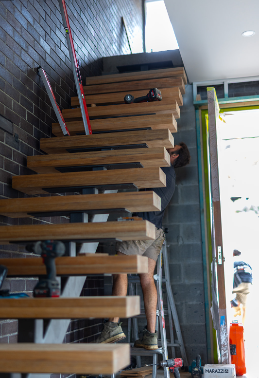 A person installing wooden stairs inside a building, using a ladder and various tools including a drill and a level.