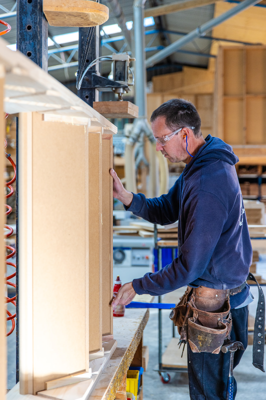 A man wearing safety glasses and a hoodie working on a woodworking project in a workshop, using glue on a wooden piece.