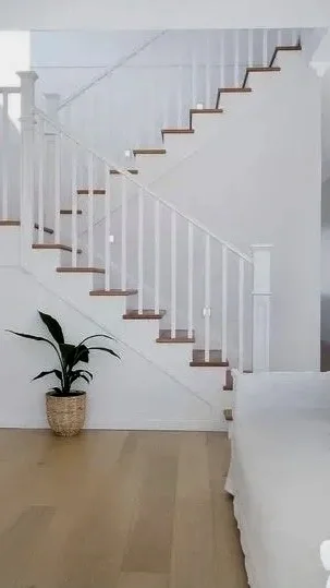 A white staircase with wooden steps and white railings inside a home, with a potted plant on the floor at the bottom of the stairs.