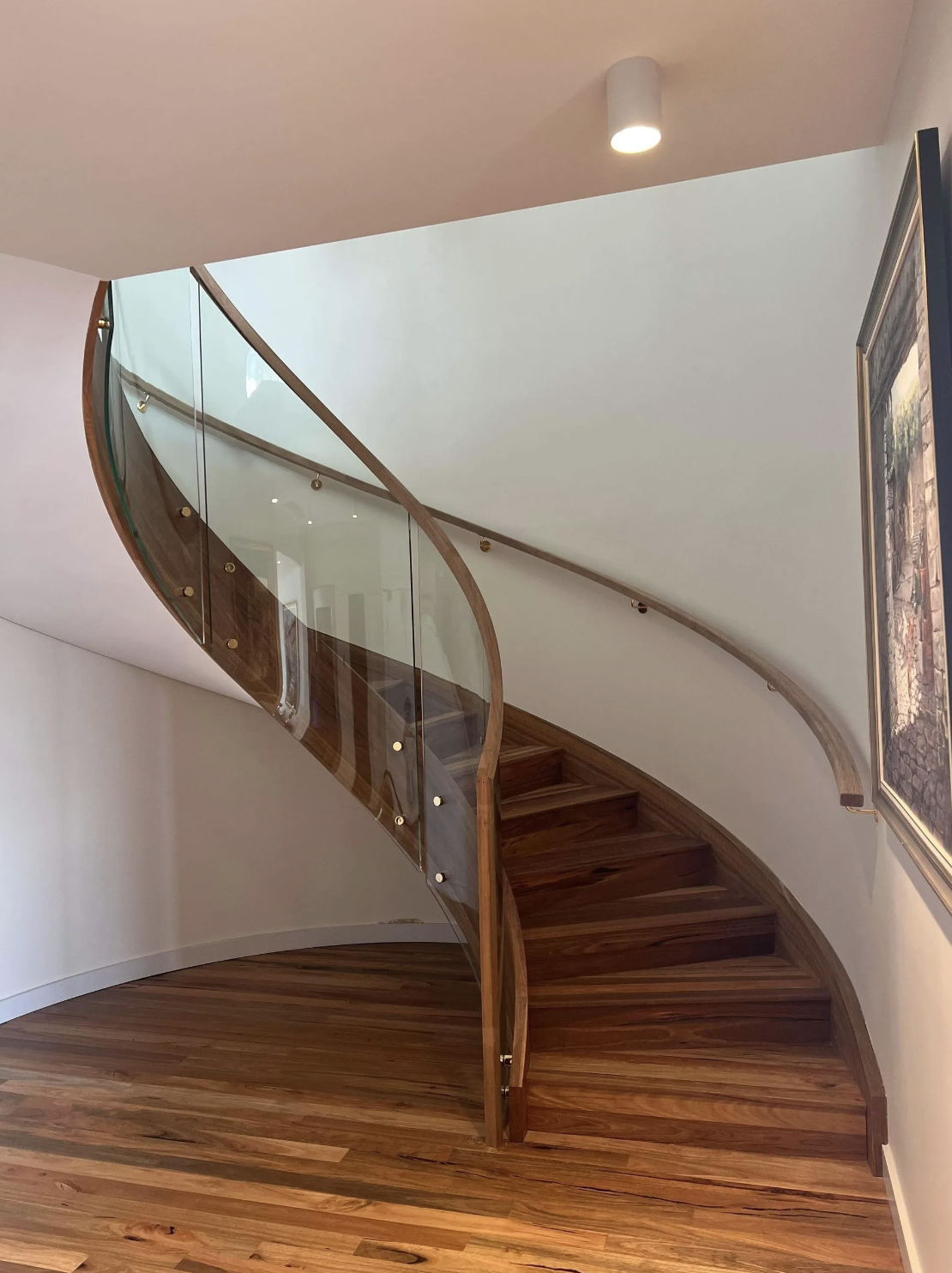 A wooden staircase with glass railing and brass fixtures in a modern interior, featuring a curved design and hardwood flooring.