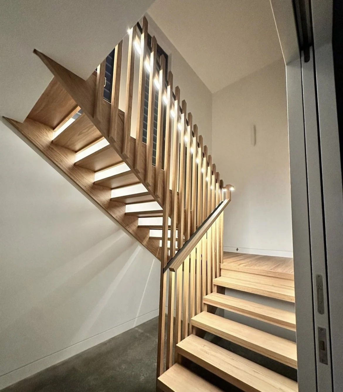 Modern wooden staircase with vertical railings and integrated lighting, viewed from the bottom staircase looking upward.