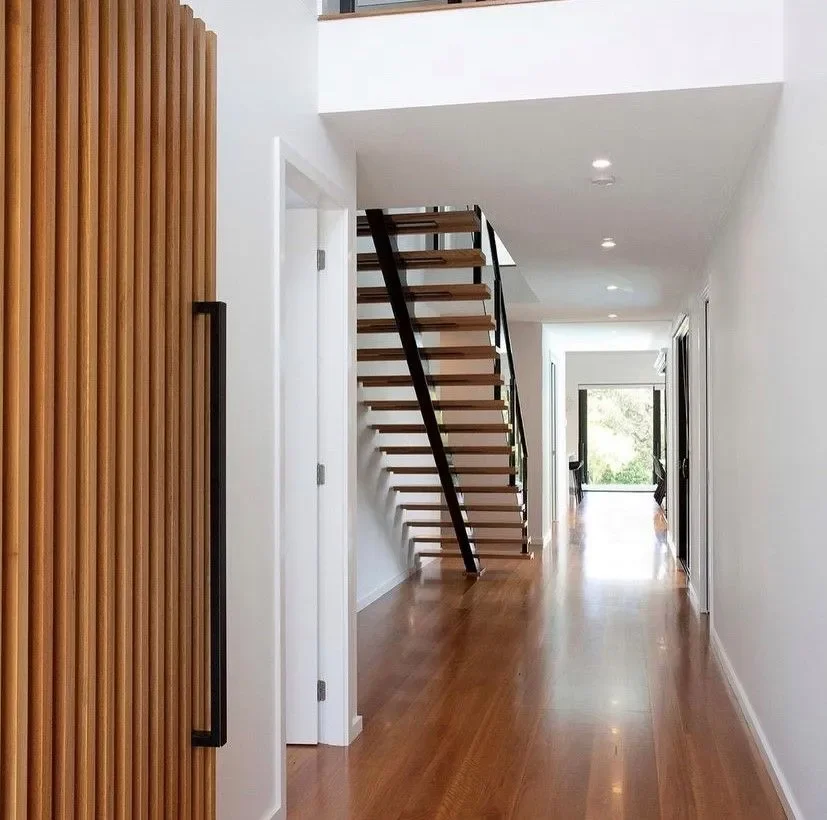 Modern hallway with wooden floors, white walls, a staircase with open risers and black railing, and a door with glass at the end letting in natural light.