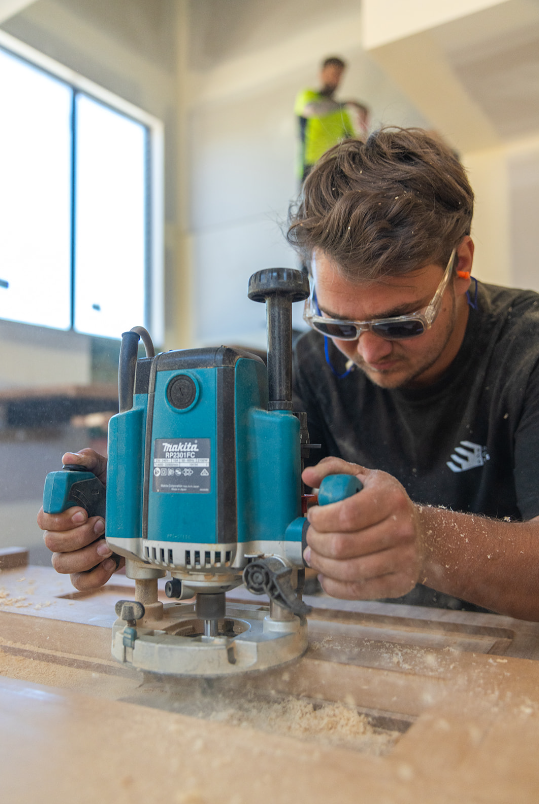 A young man with safety glasses working with a router on a piece of wood in a woodworking workshop. A man in a safety vest is standing in the background near a large window.