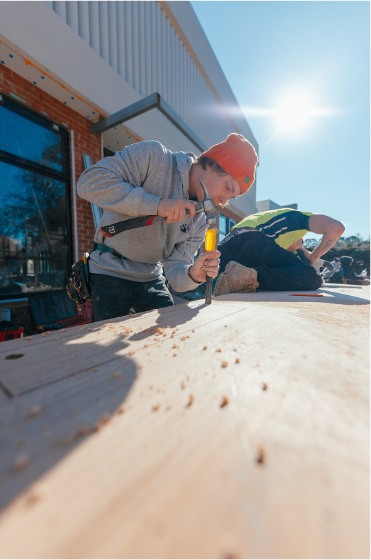 Young man wearing an orange beanie using a hammer and chisel on a large wooden surface outdoors, with another person working nearby and a modern building in the background under a bright blue sky.