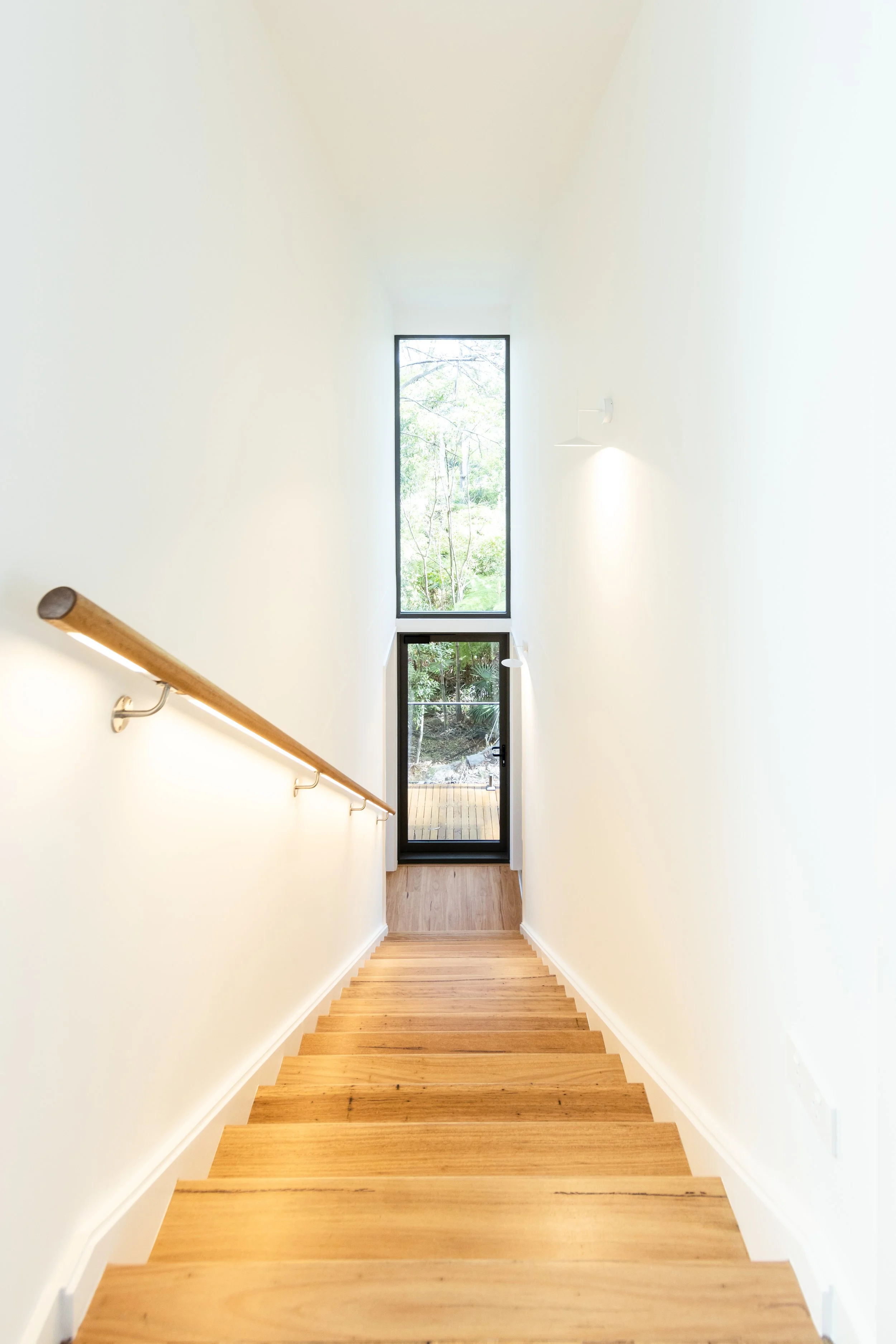View of wooden staircase leading down to a glass door with a window above, overlooking a greenery landscape outside.