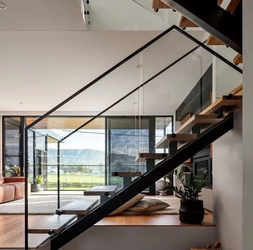 Modern living room with a glass staircase, large windows showing a green outdoor landscape, and wooden flooring.
