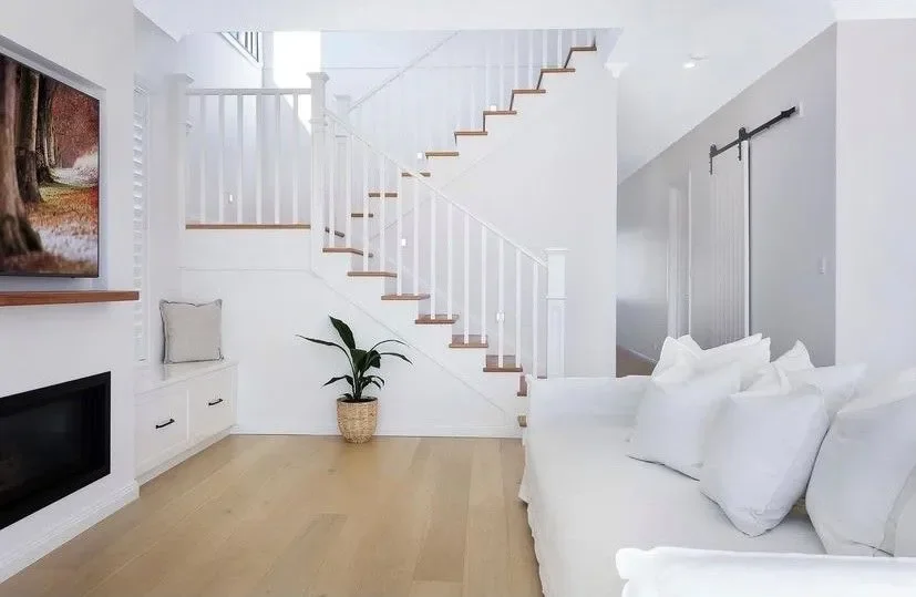 Bright living room with white walls, staircase with white railings, a fireplace, a potted plant, and a white sofa with pillows.