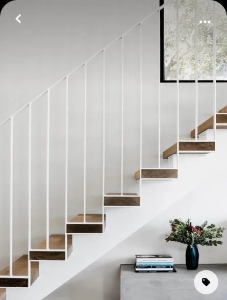 Interior of a modern staircase with wooden steps and white railings, next to a wall with a window, and a small table with a bouquet of flowers and books underneath.