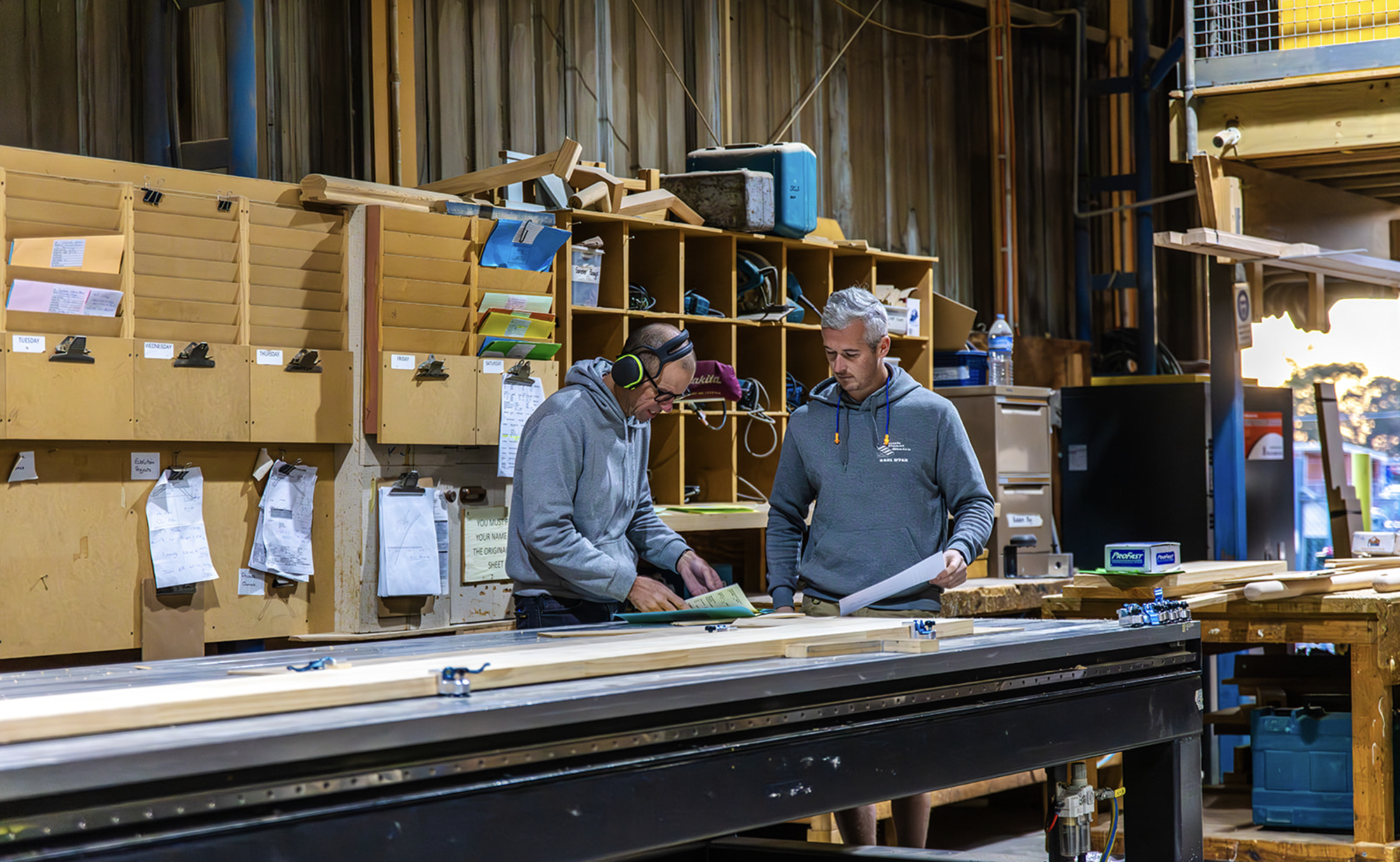 Two men in a woodworking shop examining plans, surrounded by wood, tools, and storage units.