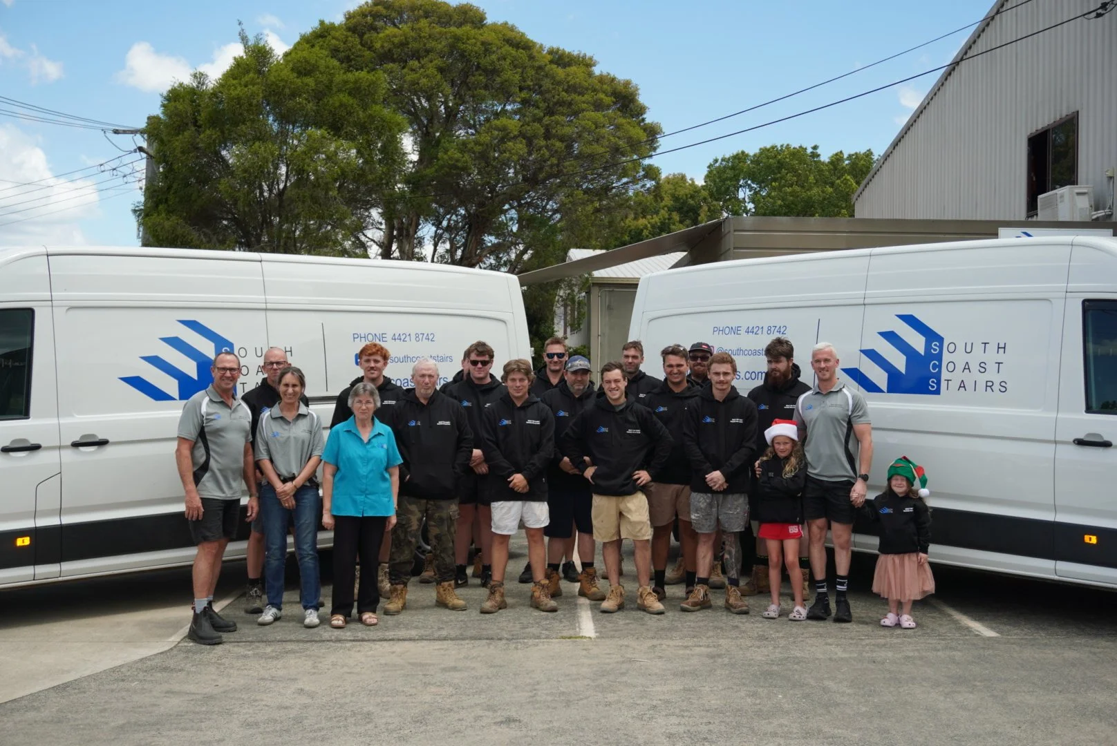 Group of people, including adults and children, standing outdoors between two white vans with South Coast Stairs logos. Some of the children are wearing Christmas hats, and the scene is set on a cloudy day with trees in the background.