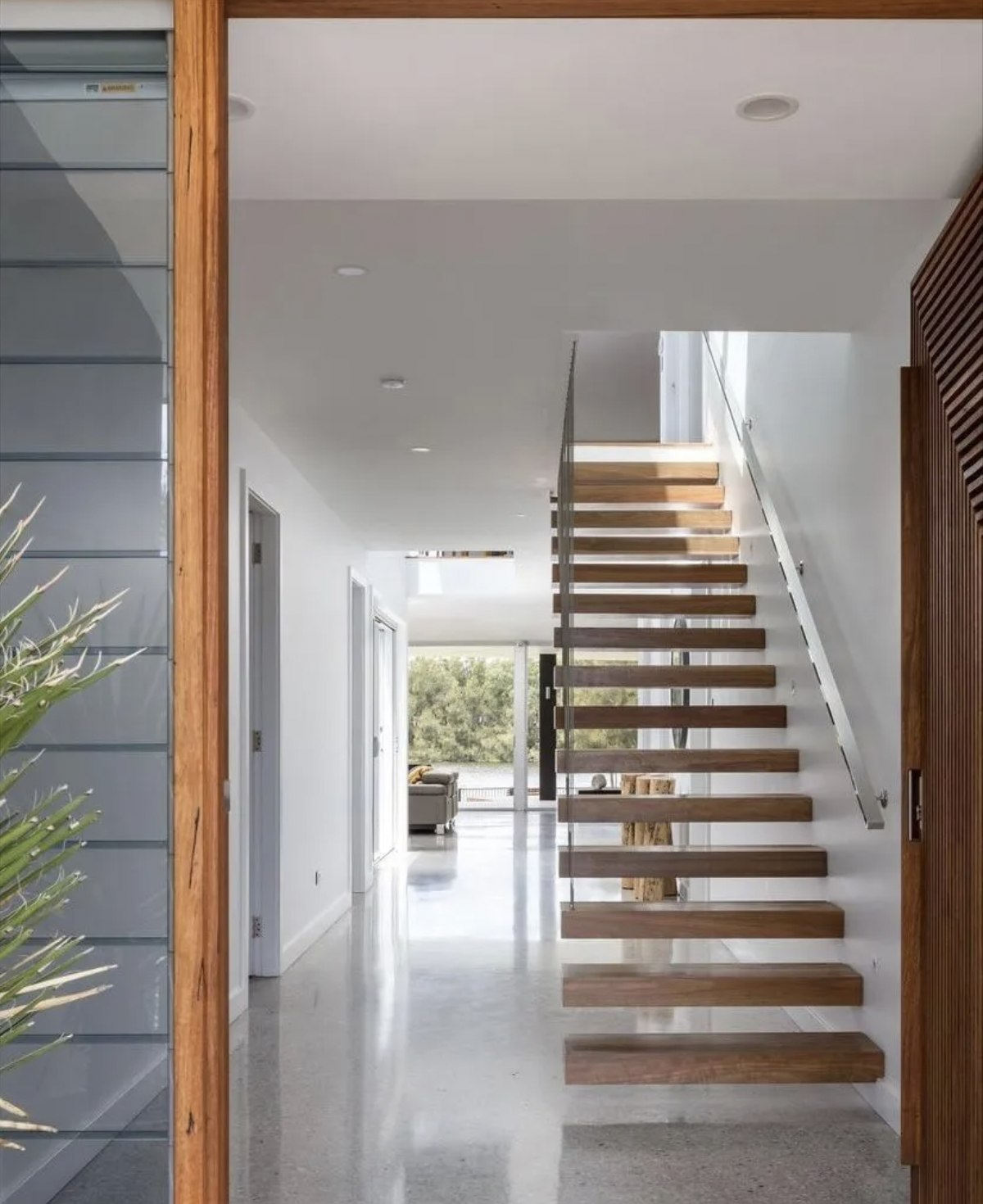 Interior of a modern, minimalist home with a floating wooden staircase, white walls, and a polished concrete floor, with a view of a living area with large windows showing trees outside.