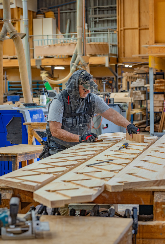 A man working on a woodworking project in a workshop, cutting and assembling wooden pieces on a large workbench surrounded by tools and wood shelves.