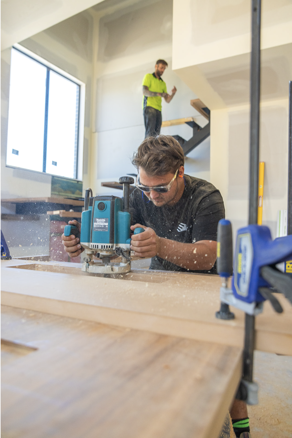 A man using a router to cut wood at a workbench in a woodworking shop, with a worker in a neon green shirt on stairs in the background.