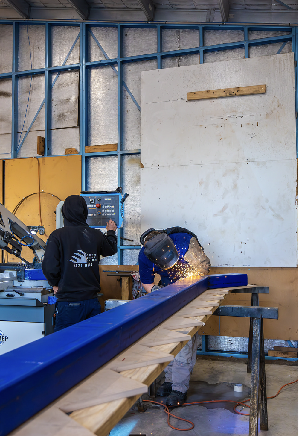Two workers in a workshop, one operating a control panel and the other welding a blue metal beam, with sparks flying.
