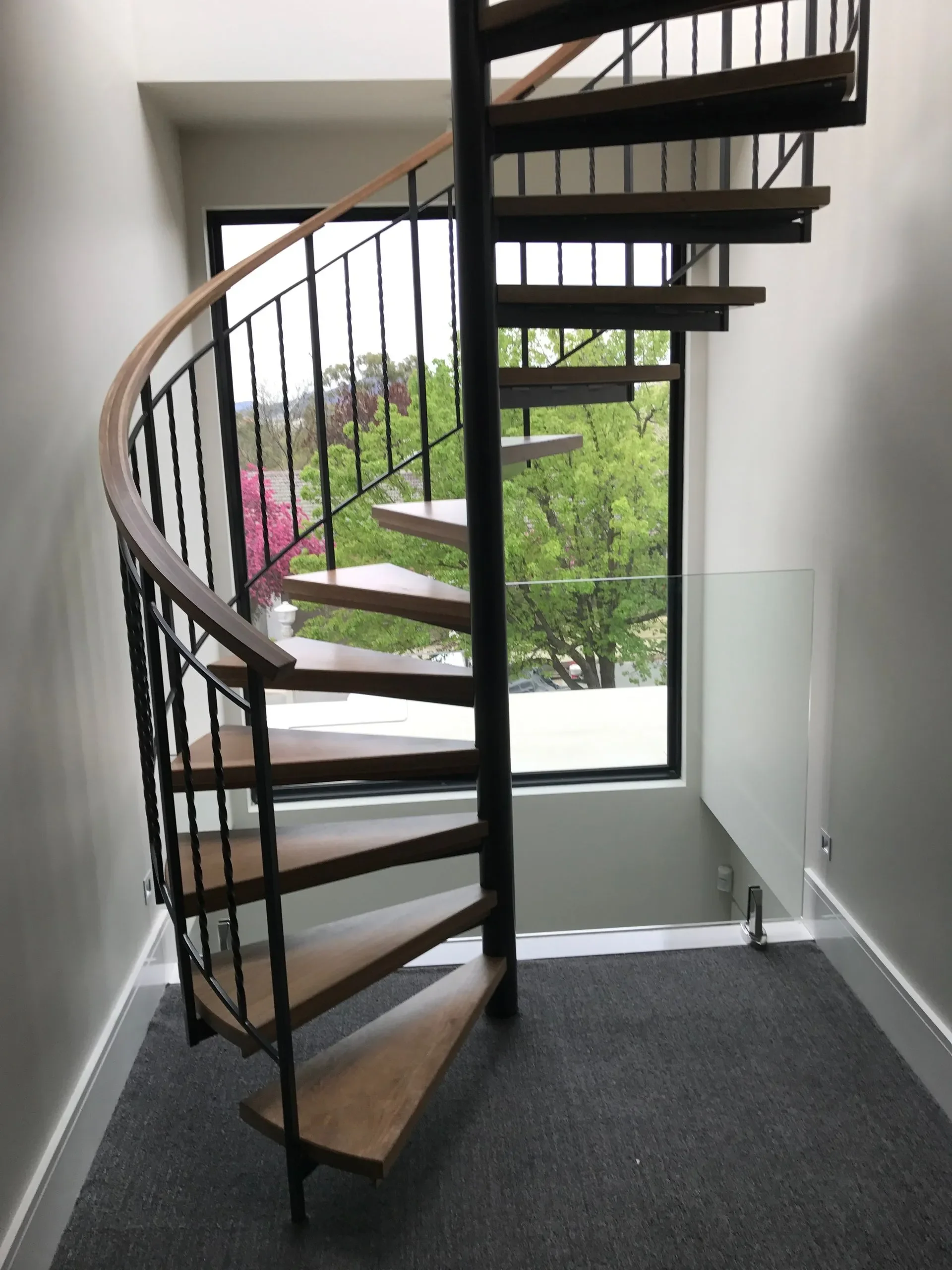 Interior view of a modern spiral staircase with wooden steps, black metal railing, next to a large window showing daylight and green trees outside.