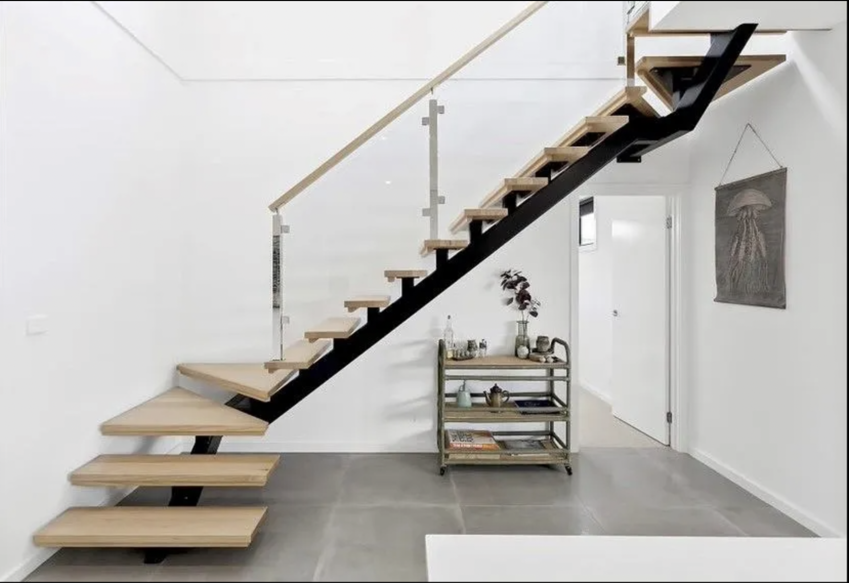 Interior of a modern house showing wooden floating stairs with black support, a metal and wood cart with decorative items and plants underneath the stairs, a white door, a wall hanging with a jellyfish design, and a small window on the wall.