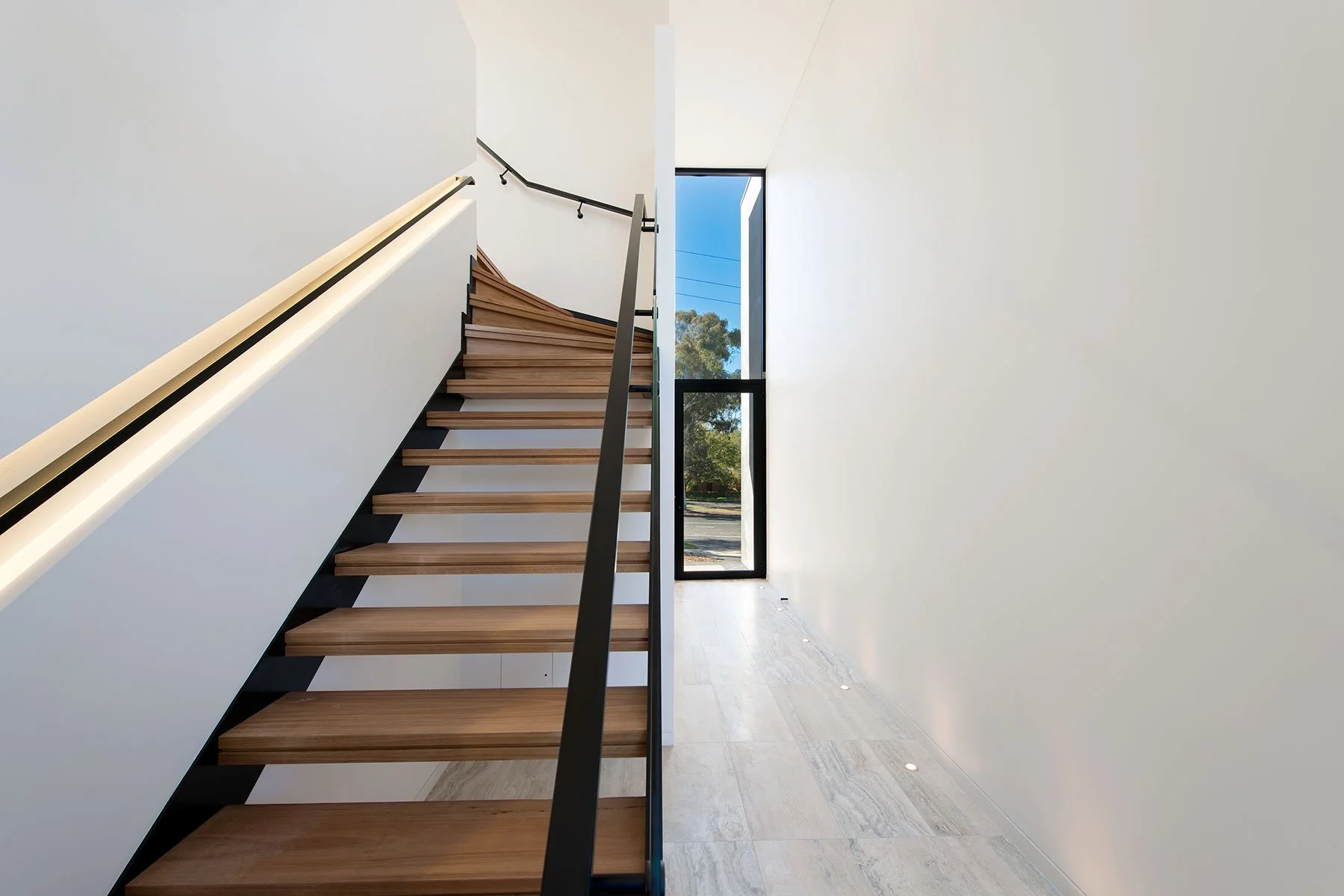 Interior view of a modern staircase with wooden steps, black handrail, and white walls, next to a large window showing trees outside.