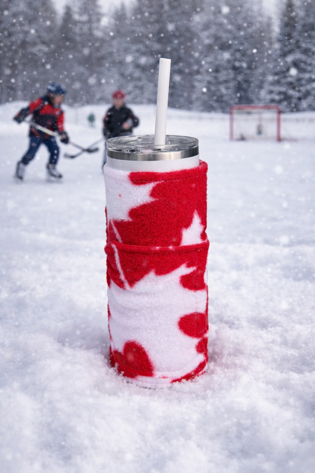 A drink in a clear tumbler with a straw, wrapped in a red and white festive cozy, sitting on snow with a hockey game and snow-covered trees in the background.