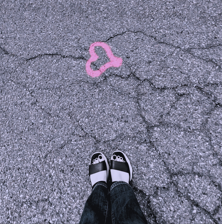 Photo showing a person's feet in black and white sandals on cracked pavement, with pink heart-graffito on the ground.