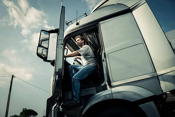 A man sitting in the driver’s seat of a semi-truck with the door open, looking out, against a cloudy sky.