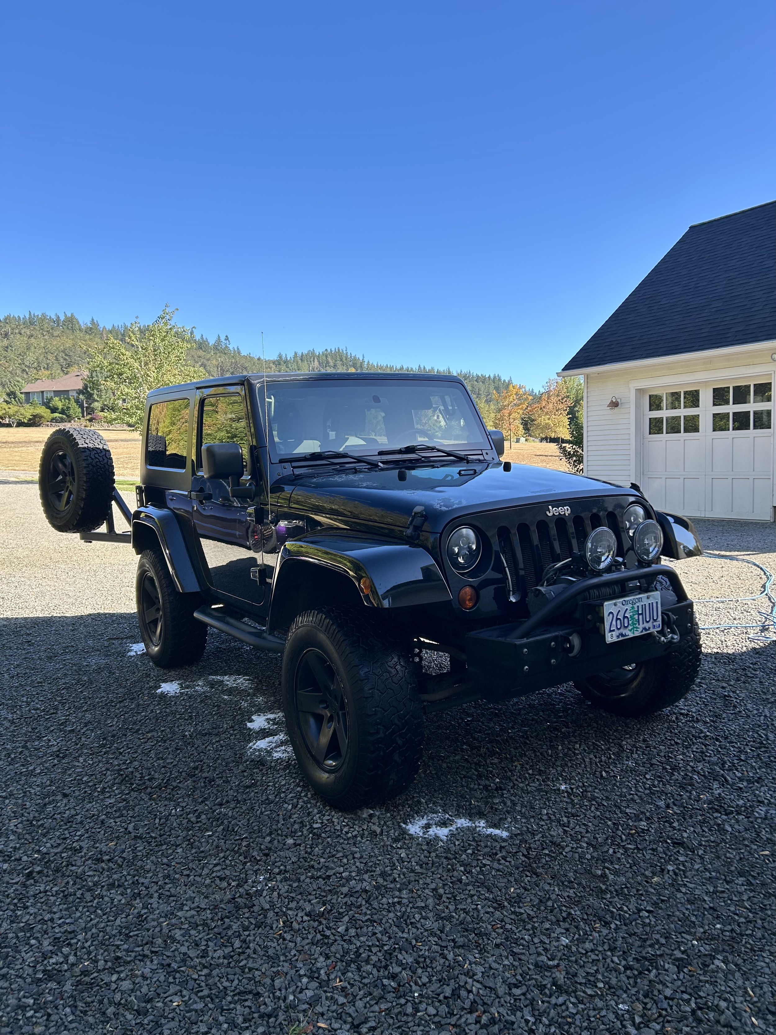 Black Jeep Wrangler parked on gravel driveway near house under blue sky with trees and hills in background.