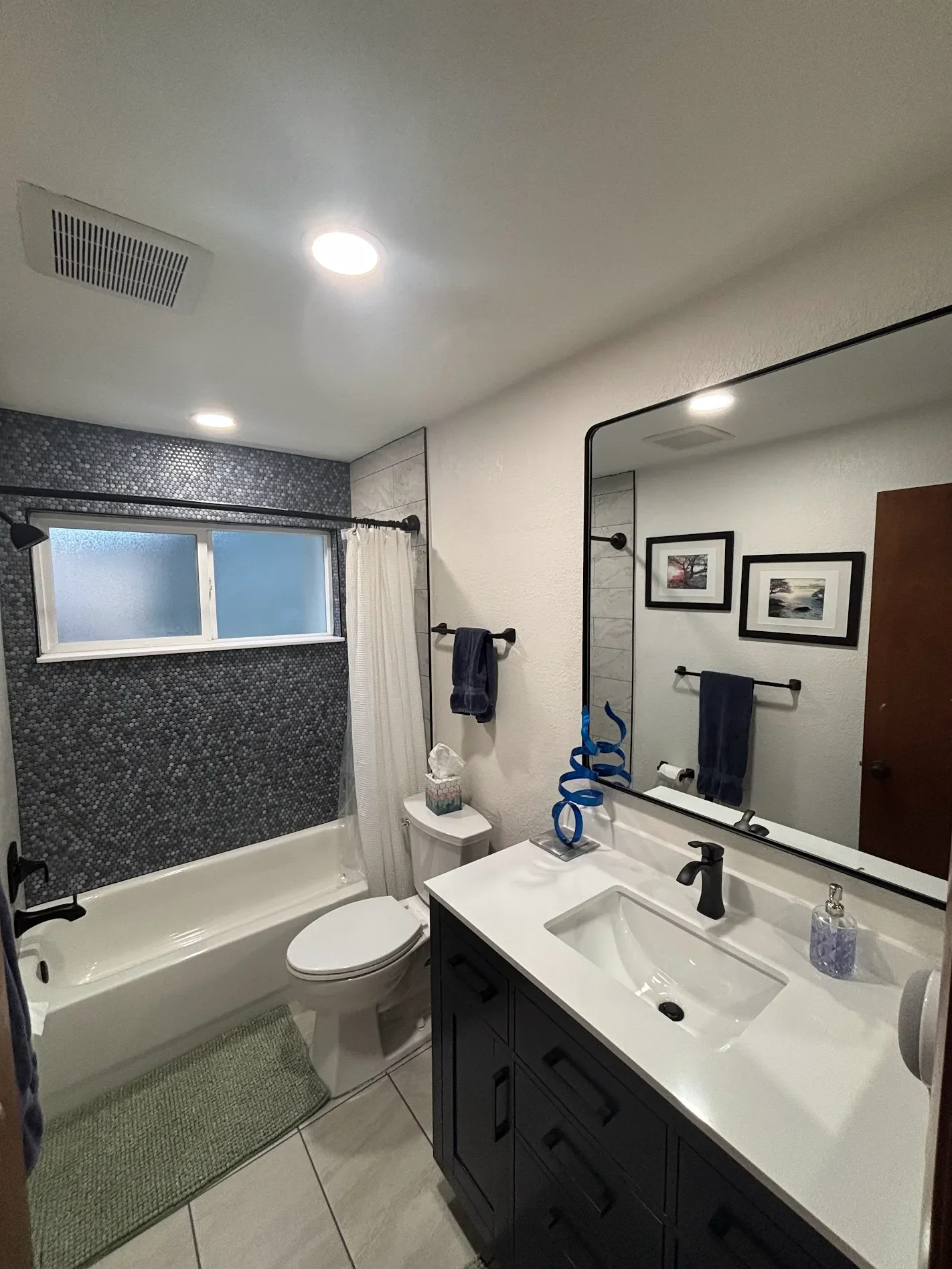 A bathroom with a shower, toilet, sink, and mirror. The shower has dark pebble-like tiles and a frosted window. The vanity has a black cabinet, white countertop, and a large rectangular mirror. Decor includes framed artwork, towels, and a decorative sculpture. Recessed ceiling lights illuminate the space.